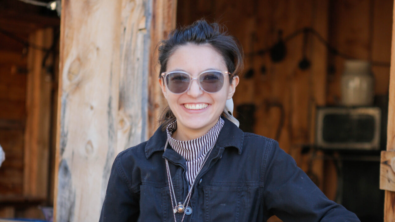 Una persona que sonríe al aire libre, con gafas de sol y una camiseta a rayas bajo una chaqueta oscura