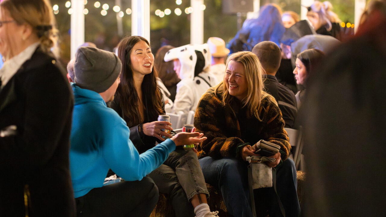 Gente charlando y riendo en un evento al aire libre con guirnaldas de luces festivas