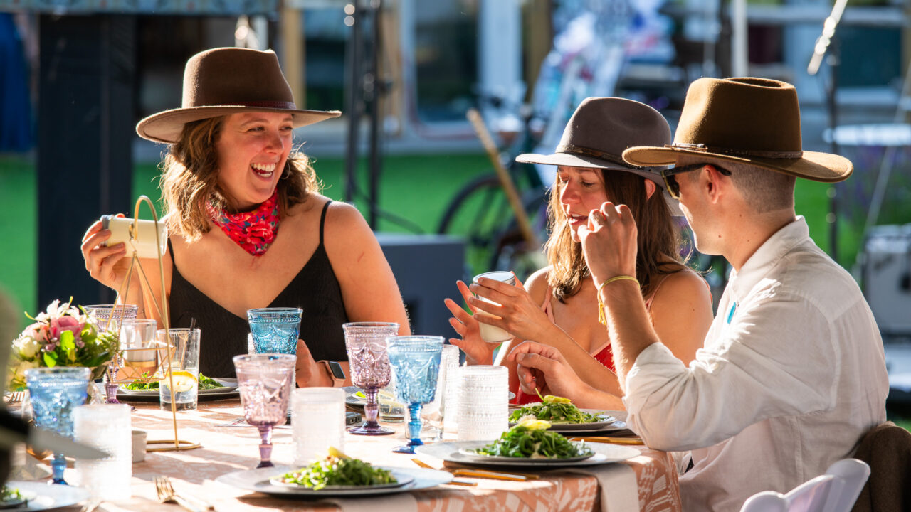 Tres personas con sombreros riendo en una mesa al aire libre, con comida y bebida