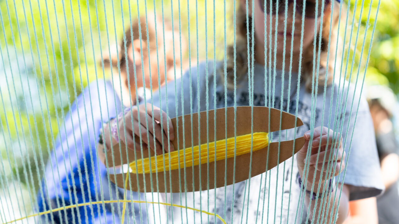 Child weaving with a loom, holding a shuttle threaded with yellow yarn